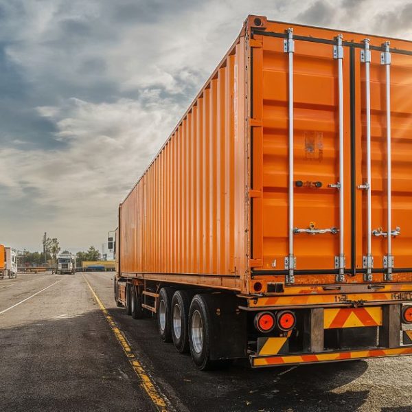 Rear view of a large semi-truck carrying a bright orange cargo container on an asphalt lot under a sunny sky.