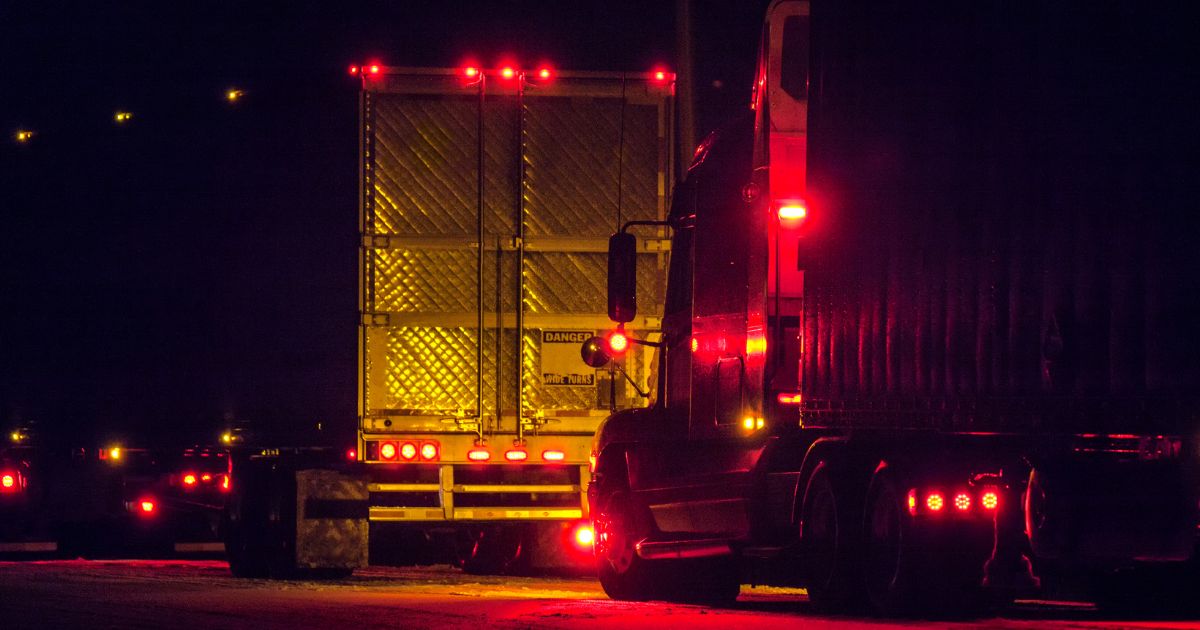 Nighttime view of semi-trucks on a dark road with bright red taillights and clearance lights illuminated.